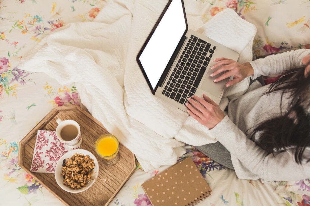 Kobieta pracuje na laptopie w łóżku z tacą ze śniadaniem.